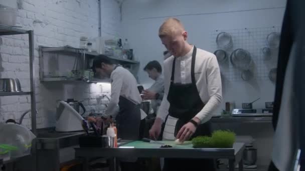 エプロンの男は台所の机のそばに立って野菜を切る バックグラウンドで美食料理を作るプロの料理人のグループ シェフは仕事のプロセスを制御します レストランでのチームワークの概念 — ストック動画