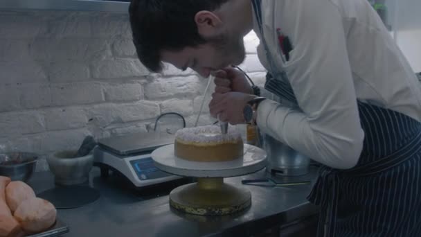 エプロンのプロのお菓子は パイプバッグでホイップクリームを絞り グルメケーキの最終的なタッチを作ります 背景にある製品や調理ユーティリティ レストランキッチンで働くことのコンセプト — ストック動画