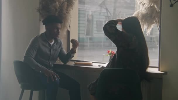 アフリカ系アメリカ人男性が窓の近くのテーブルに座り 女性と物語を語り合います カフェでのデートで複数の民族のカップル モダンなデザインのレストランでお客様 一般食の概念 — ストック動画
