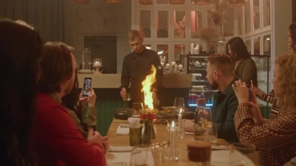 男性シェフは 現代のガストロカフェの人々の前で炎のスタイルで食事を準備します 彼らは携帯電話でプロセスを撮影します 友人のグループは レストランで週末の夜を過ごす 一般食の概念 — ストック動画