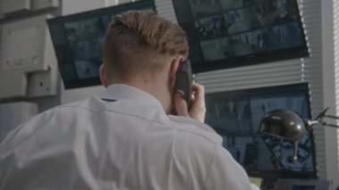 Güvenlik operatörü gözlem odasında sabit hatlı telefonla konuşuyor. Güvenlik görevlisi bilgisayardaki CCTV kameralarını izliyor. Ekranlarda yüz tanıma yazılımlı güvenlik kameraları görülüyor. Arka plan.