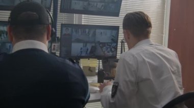 Kulaklıklı güvenlik görevlisi bilgisayardaki kameraları izliyor. Santral sabit telefon ve telsiz kullanıyor. Yapay zeka yüz tarama programıyla CCTV kameralarını gösteren birden fazla ekran var. Arka plan.