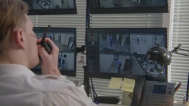 Güvenlik görevlisi ofis kameralarını bilgisayardan izliyor, telsiz kullanıyor. Yüksek teknolojili yazılım, ekranlarda yüz tanıma sistemi olan güvenlik kameralarını gösteriyor. Sosyal güvenlik kavramı.