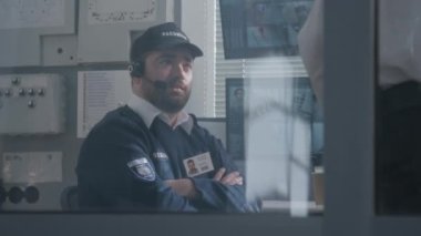 Kulaklıklı güvenlik görevlisi iş arkadaşıyla konuşuyor. Erkekler gözetleme odasında çalışıyor. Bilgisayar monitörlerinde gösterilen güvenlik kameralarının görüntüsü. Gözlem ve CCTV teknolojisi. Sosyal güvenlik kavramı.