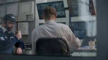 Güvenlik görevlisi, iş arkadaşıyla birlikte CCTV kameralarını izlerken kahve içiyor. Güvenlik görevlisi telsiz ve sabit telefon kullanıyor. Programdaki bilgisayar ekranlarında gösterilen güvenlik kameralarından görüntüle.