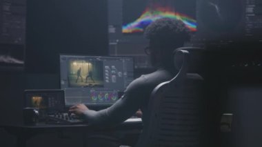 Afrikalı Amerikalı film editörü modern stüdyoda bilgisayar üzerinden renk değerlendirmesi yapıyor. Duvarda RGB renk düzeltme çubuğu ve program arayüzü olan büyük monitörler. Video prodüksiyonu. El bilgisayarı.