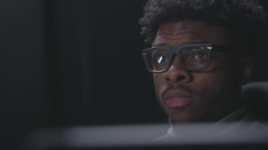 Modern tasarımla stüdyoda çalışan ya da film izleyen Afro-Amerikan bir adamı yakından çek. Film editörü monitörlere bakar. Video görüntüleri gözlüklere yansıyor. Serbest ve uzaktan kumandalı. Yakınlaştır.