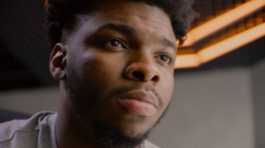 Genç Afro-Amerikan bir adamın detaylı portresi. Ciddi bir yüz, kendine güvenen ve odaklanmış bir görünüm. Yoğunlaşmış adam başka tarafa bakıyor, gözlerini kırpıyor, kameraya poz veriyor. Adam stüdyoda duruyor. Arka planda LED lambaları.