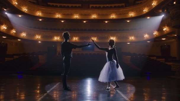 古典的なバレエダンサーのカップルは 公演前に劇場舞台で練習します トレーニングスーツの男は優雅なバレリーナを持ち上げる ダンス振付稽古 照明付きの劇場ホール スローモーション — ストック動画