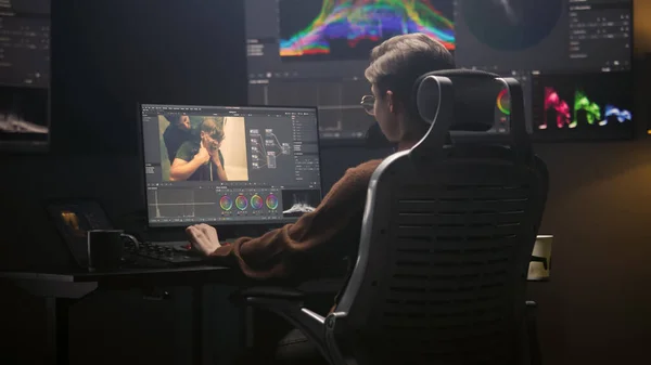 Female editor works in modern studio on computer using professional editing software. Woman edits video footage, makes color grading for movie post production. Big screen with RGB graphic on the wall.