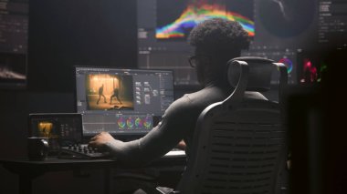 Afrikalı Amerikalı film editörü modern stüdyoda bilgisayar üzerinden renk değerlendirmesi yapıyor. Duvarda RGB renk düzeltme çubuğu ve program arayüzü olan büyük monitörler. Video prodüksiyonu. El bilgisayarı.