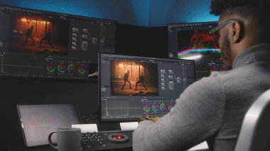 Afrikalı Amerikalı renkçi modern stüdyoda kontrol makinesinde renk değerlendirmesi yapıyor. Hareketli film görüntüleri ve duvarında RGB renk düzeltme çubuğu olan büyük ekranlar. Film sonrası üretim süreci.