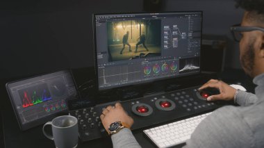 Afrikalı Amerikalı renkçi modern stüdyoda kontrol makinesinde renk değerlendirmesi yapıyor. Hareketli film görüntüleri ve duvarında RGB renk düzeltme çubuğu olan büyük ekranlar. Film sonrası üretim süreci.
