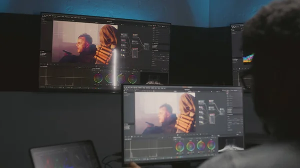 African American movie maker does color grading on computer in studio with equipment. Big monitors with action film footage and professional software interface on wall. Video post production process.