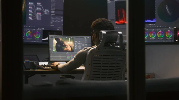 Experienced African American video maker does color grading in program on computer. Process of colour correction for movie post production in modern studio. Multiple monitors with action film footage.