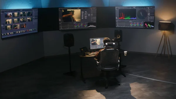 The video editor works in studio on computer, uses color grading control panel, edits video, makes color correction for movie. Big screens showing software interface with film footage and RGB graphic.