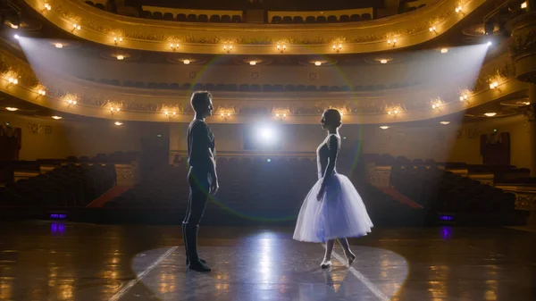 Couple of classical ballet dancers practice on theatre stage before performance. Man in training suit lifts graceful ballerina. Dance choreography rehearsal. Illuminated theatrical hall. Slow motion.