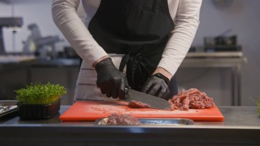 Önlüklü profesyonel aşçı kesim tahtasındaki et parçalarını kesiyor. Mutfak masasında bir kutu mikroçim duruyor. Meslektaşlar arka planda çalışır. Şefin elleri yakın plan. Kamu hizmeti. Gastronomik sanat.