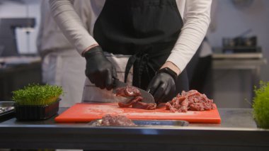 Önlüklü profesyonel aşçı kesim tahtasındaki et parçalarını kesiyor. Mutfak masasında bir kutu mikroçim duruyor. Meslektaşlar arka planda çalışır. Şefin elleri yakın plan. Kamu hizmeti. Gastronomik sanat.