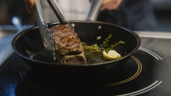 stock image The chef cooks delicious meal on stove. The cook fries piece of juicy pork meat on frying pan with rosemary, pours oil using spoon. Process of steak cooking. Restaurant with professional kitchen.