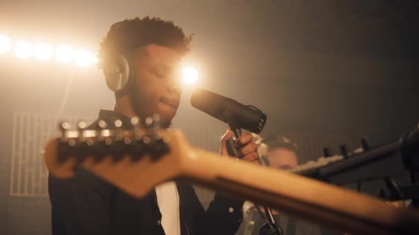 非裔美国歌手在隔音室内对着话筒唱着新的曲子 摇滚乐队在录音室排练 男人在背景音乐中弹吉他 音乐制作的概念 — 图库视频影像