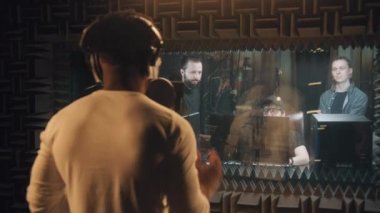 Afrikalı Amerikalı şarkıcı dans ediyor ve ses kayıt stüdyosunda mikrofona yeni bir şarkı söylüyor. Müzik grubu ve deneyimli ses mühendisi vokalistlere bakar. Müzik üretimi kavramı. Arka plan.