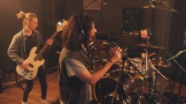 Profesyonel kulaklıklı vokalist ses geçirmez odada mikrofona beste yapıyor. Müzisyenler gitar ve davul setiyle çalarlar. Ses kayıt stüdyosundaki havalı rock grubu. Müzik üretimi kavramı.