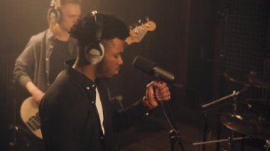 Afrikalı Amerikalı şarkıcı jestler yapıyor ve ses geçirmez odada mikrofona şarkı söylüyor. Adam arka planda elektro gitarla çalıyor. Ses kayıt stüdyosunda prova. Müzik üretimi.