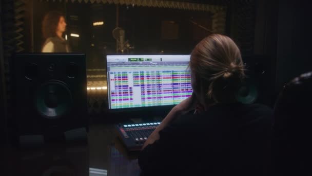 経験豊富な女性サウンドエンジニアは オーディオ録音スタジオで歌手と仕事をします 男は防音室でマイクに作曲を歌う プロのプログラムを示すコンピュータ画面 音楽制作 — ストック動画