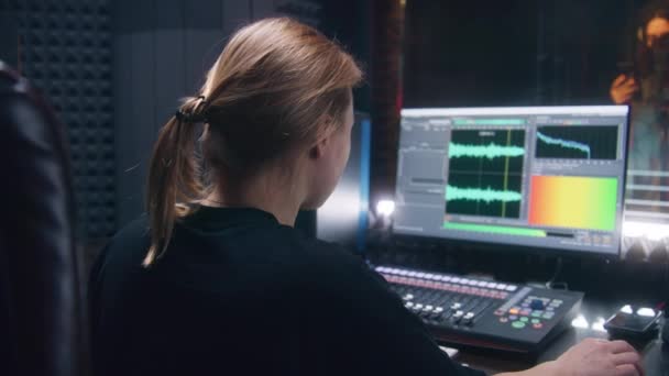 女性サウンドエンジニアはコントロールミキシングサーフェスを使用します 音楽を作成するためのDawソフトウェアインターフェースを示すコンピュータ画面 シンガーは防音室で歌を録音します 録音スタジオだ 音楽制作 — ストック動画