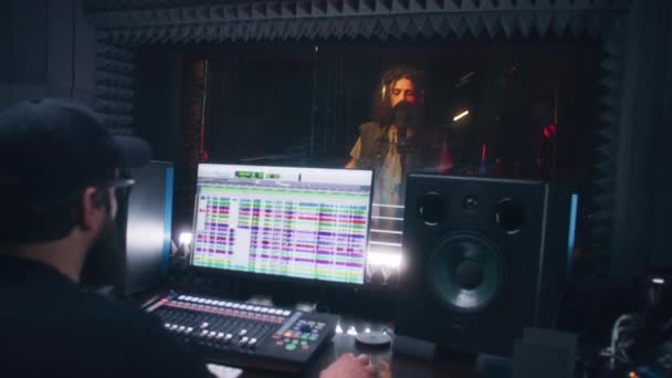 男性歌手は防音室で歌を歌う オーディオエンジニア プロデューサーは コンピュータ上で音楽を作成するためのサウンドトラックやツールを持つプロのDawソフトウェアを使用しています 録音スタジオだ 音楽制作 — ストック動画