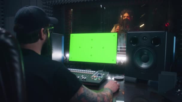 男性歌手は防音室で歌を録音します サウンドエンジニアはコントロールミキシングサーフェスを使用します コンピューター画面には Dawソフトウェアインターフェースと音楽制作のためのツールが表示されます 録音スタジオだ 緑の画面 — ストック動画