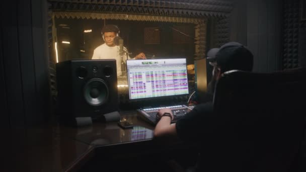 プロフェッショナルヘッドフォンのアフリカ系アメリカ人男性は 録音スタジオでマイクに新しい曲を歌います サウンドエンジニアは デジタルオーディオワークステーションソフトウェアによってボーカルを記録します 音楽制作の概念 — ストック動画