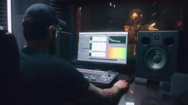 Erkek şarkıcı ses geçirmez odada şarkı kaydediyor. Ses mühendisi, yapımcı kontrol yüzeyini kullanıyor. Müzik yaratmak için DAW yazılım arayüzünü gösteren bilgisayar ekranı. Ses kayıt stüdyosu. Müzik üretimi.