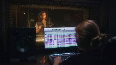 Ses mühendisi ses kayıt stüdyosunda şarkıcıyla çalışıyor. Kulaklıklı vokalist ses geçirmez odada şarkı söylüyor. Bilgisayar ekranı, şarkı çalma parçalarıyla müzik yapmak için DAW yazılım arayüzünü gösterir.