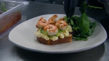 Şef yemek sunumu yapıyor. Aşçı marul ve domatesleri kızarmış yumurta, avokado, kızarmış karides ve sos ile birlikte tabağa koyar. Profesyonel mutfağı olan bir restoran. Kamu hizmeti.