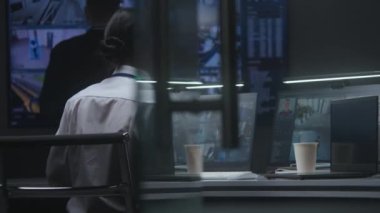 Kadın güvenlik görevlisi, polis izleme merkezinde bilgisayar üzerinde çalışıyor. Güvenlik kameralarını yapay zeka yüz tanıma sistemiyle kontrol ediyor. Erkek işçi, güvenlik kamerası görüntüleriyle büyük dijital ekrana bakıyor..