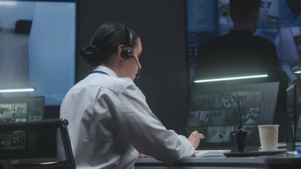 戴耳机的女警官在电脑上工作 在警察监控中心用闭路电视摄像头录像 在收音机里交谈 男性员工在大屏幕上控制监控摄像头 — 图库视频影像