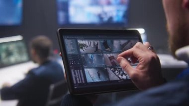 Güvenlik görevlisinin güvenlik kameralarını AI yüz taramasıyla yakınlaştırırken polis gözetleme merkezinde tablet kullanırken yakın plan görüntüsü. Meslektaşlar arka planda çalışır. İzleme sistemi. Sosyal güvenlik kavramı.