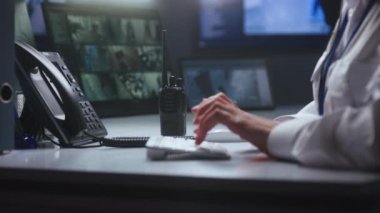 Kadın güvenlik görevlisi polis gözlem merkezinde çalışıyor. Klavyede kadın tipi var, sabit telefon ve radyo kullanıyor. Güvenlik kameraları bilgisayar ve tablet ekranlarda görünüyor. Kapat..