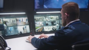 Erkek güvenlik operatörü AI yüz taraması ve telsizle konuşmalarla CCTV kameralarını kontrol ediyor. Bilgisayar monitörleri güvenlik kamerası görüntülerini gösteriyor. İzleme ve izleme sistemleri. Sosyal güvenlik kavramı.