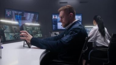 Erkek güvenlik görevlisi tablete dokunuyor ve yapay zeka yüz taramasıyla mobese kameralarını kontrol ediyor. Bilgisayar monitörleri ve güvenlik kamerası görüntülerini gösteren büyük dijital ekranlar. Bayan meslektaşım arka planda çalışıyor..