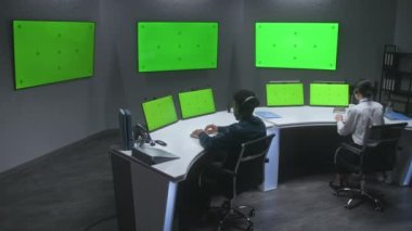 Çok ırklı erkek ve kadın memurlar CCTV kameralarını kontrol ediyor, güvenlik kontrol merkezinde çalışıyorlar. Büyük dijital ekranlar ve yeşil ekranlı bilgisayar monitörleri, güvenlik kameralarının görüntülerini gösteriyor..
