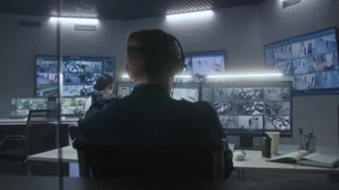 Çeşitli polis ekipleri yapay zeka yüz tanıma sistemine sahip CCTV kameralarını izliyor. Çalışanlar güvenlik kontrol merkezinde çalışıyor. Bilgisayar monitörleri ve güvenlik kameralarının görüntülerini gösteren büyük dijital ekranlar..