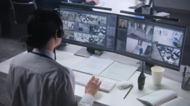 Kulaklıktaki güvenlik görevlisi, bilgisayarında yapay zeka yüz tanıma sistemi olan kameraları kontrol ediyor. Kadın çalışan güvenlik kontrol merkezinde çalışıyor. Bilgisayar monitörleri güvenlik kamerası görüntülerini gösteriyor.