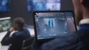 Güvenlik operatörünün güvenlik kameralarını AI yüz tanıma sistemiyle yakınlaştırırken polis gözetleme merkezinde tablet kullanırken yakın plan görüntüsü. Meslektaşlar arka planda çalışır. İzleme sistemi. Sosyal güvenlik kavramı.