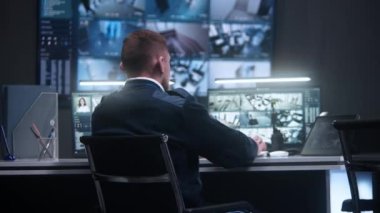 Erkek memur, yapay zeka yüz taramasıyla güvenlik kameralarını kontrol ediyor. Kadın operatör polis gözetim merkezinde çalışıyor. Duvardaki büyük dijital ekranlar ve CCTV kameralı bilgisayar monitörleri. Arka plan.