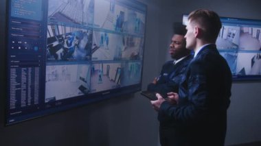 Güvenlik operatörü, güvenlik kameralarını tablet kullanarak kontrol ediyor ve polis izleme merkezindeki Afrikalı Amerikalı iş arkadaşıyla konuşuyor. Duvardaki büyük dijital ekranlar güvenlik kamerası görüntülerini gösteriyor. Sosyal Güvenlik.