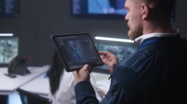 Erkek memur güvenlik kameralarını zumluyor ve kontrol ediyor. Polis izleme merkezinde tablet kullanıyor. Arka planda bilgisayar başında çalışan güvenlik görevlileri. İzleme ve izleme. Güvenlik kavramı. Kapat..