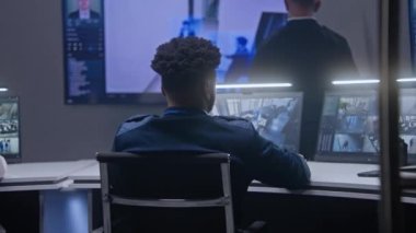 Afrika kökenli Amerikalı güvenlik görevlisi yüz tanıma sistemiyle mobese kameralarını kontrol ediyor. Adam izleme odasında iş arkadaşıyla konuşuyor. Duvarda güvenlik kameralarının görüntüsünü gösteren çok sayıda büyük ekran var. Arka plan.